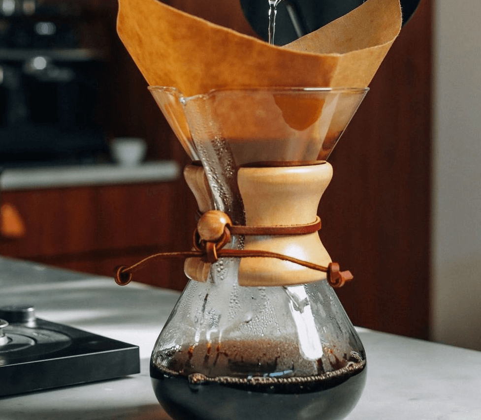 Imagem de uma jarra de vidro com coador artesanal coando café, com água sendo despejada.